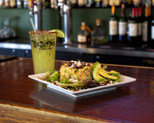 A vibrant plate of salad with avocado and a drink on a bar counter, with bottles in the background.